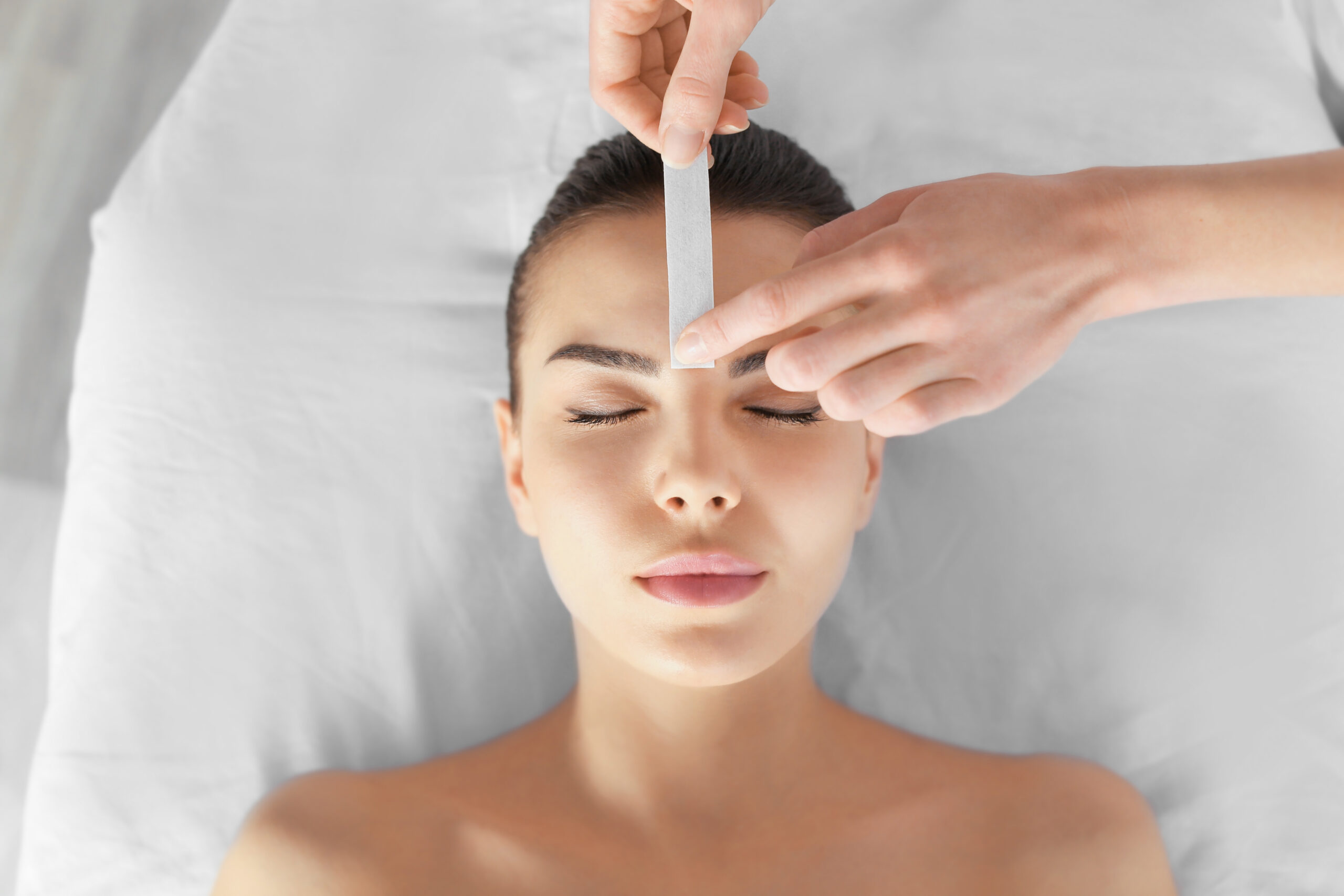 Beautician waxing young woman's eyebrows in spa center